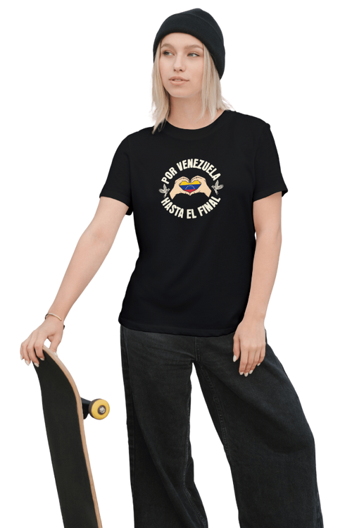 A blonde woman is wearing a black unisex t-shirt featuring a Venezuelan flag inside heart-shaped hands, the text "Por Venezuela Hasta El Final", and a pair of macaws on either side. She's holding a skateboard.