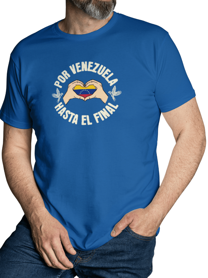 A man is wearing a blue unisex t-shirt featuring a Venezuelan flag inside heart-shaped hands, the text "Por Venezuela Hasta El Final", and a pair of macaws on either side. He stands with his left hand in his pocket.