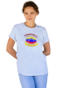 A young woman is wearing a light-blue unisex t-shirt featuring a colorful, elliptically shaped Venezuelan flag dotted (like an explosion), with the text "Venezuela Camino a La Libertad" at the top and "Hasta El Final" on a yellow ribbon below the flag. She’s standing facing forward with both hands behind her.