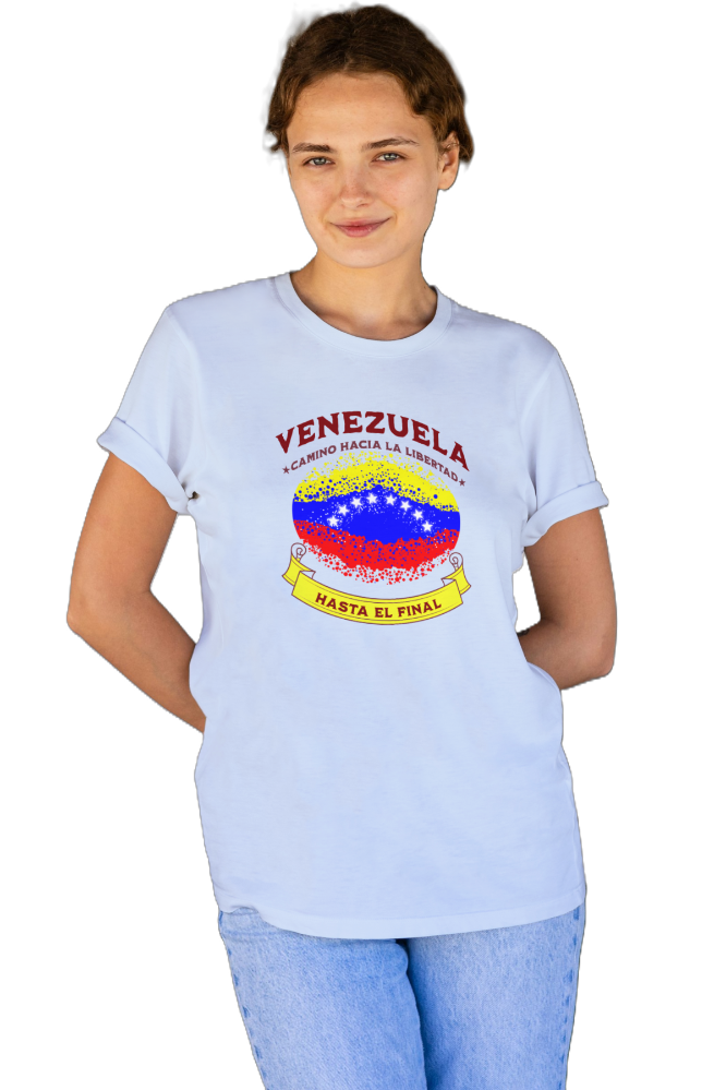 A young woman wears a light blue unisex t-shirt featuring a colorful, elliptically shaped Venezuelan flag dotted (like an explosion), with the text "Venezuela Camino Hacia La Libertad" at the top and "Hasta El Final" on a yellow ribbon below the flag.