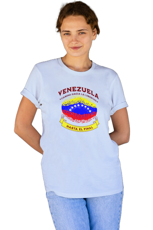 A young woman wears a light blue unisex t-shirt featuring a colorful, elliptically shaped Venezuelan flag dotted (like an explosion), with the text "Venezuela Camino Hacia La Libertad" at the top and "Hasta El Final" on a yellow ribbon below the flag.