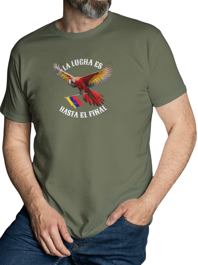 A man is wearing a military-green unisex t-shirt featuring a colorful macaw flying while holding the Venezuelan flag. The text "La Lucha Es" is at the top, and "Hasta El Final" is below the macaw. He's standing, his left hand in his pocket.