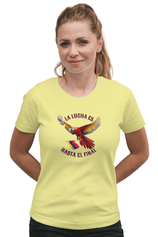 A blond woman is wearing a pale-yellow unisex t-shirt featuring a colorful macaw flying while holding the Venezuelan flag. The text "La Lucha Es" is at the top, and "Hasta El Final" is below the macaw. She's smiling, both hands behind her back.