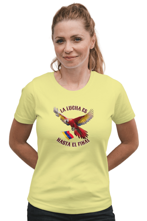 A blond woman is wearing a pale-yellow unisex t-shirt featuring a colorful macaw flying while holding the Venezuelan flag. The text "La Lucha Es" is at the top, and "Hasta El Final" is below the macaw. She's smiling, both hands behind her back.