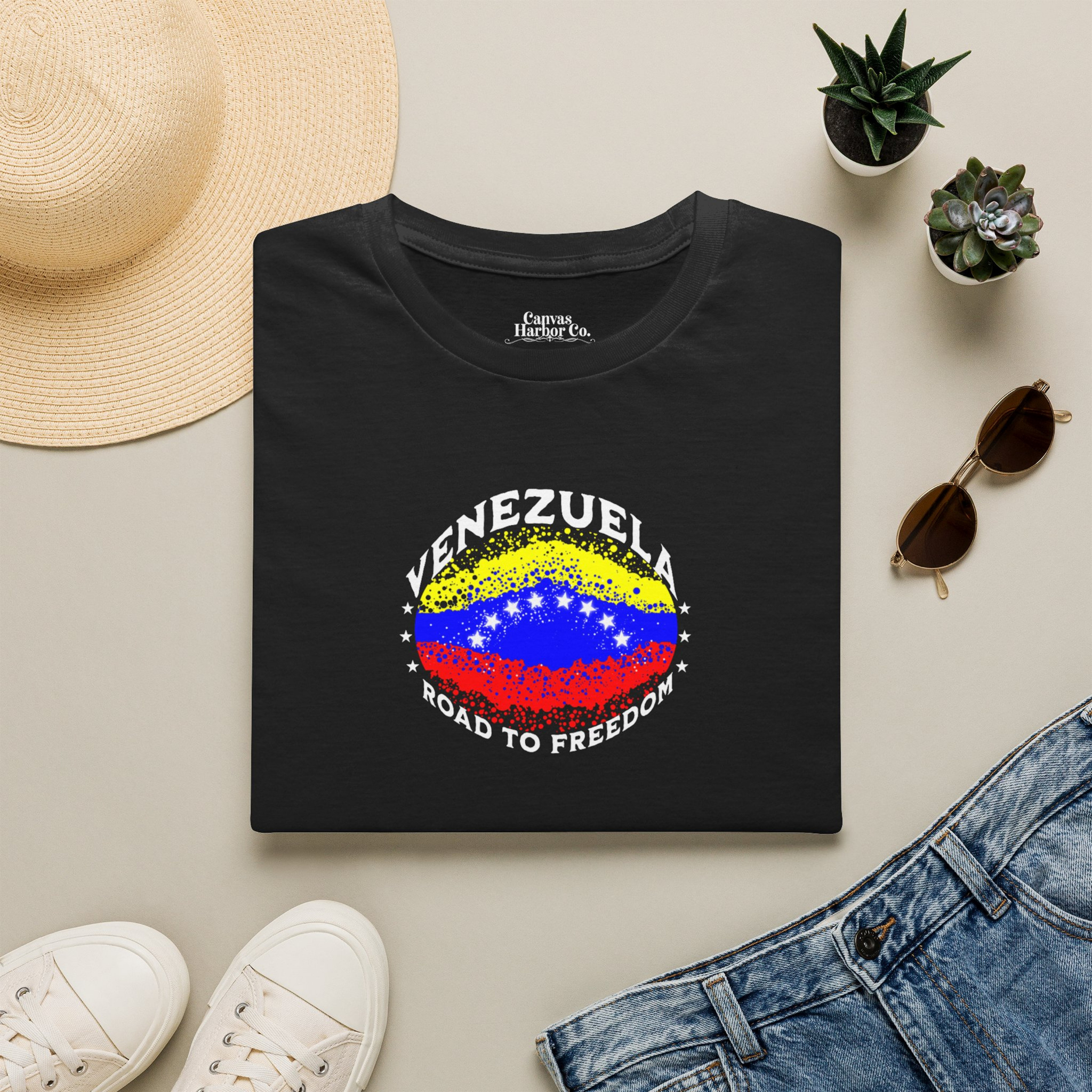 A black folded T-shirt lies on a table, featuring an elliptical Venezuelan flag dotted with dots (like an explosion), with the word "VENEZUELA” at the top, the text “ROAD TO FREEDOM” below the flag, and 3 stars on each side.