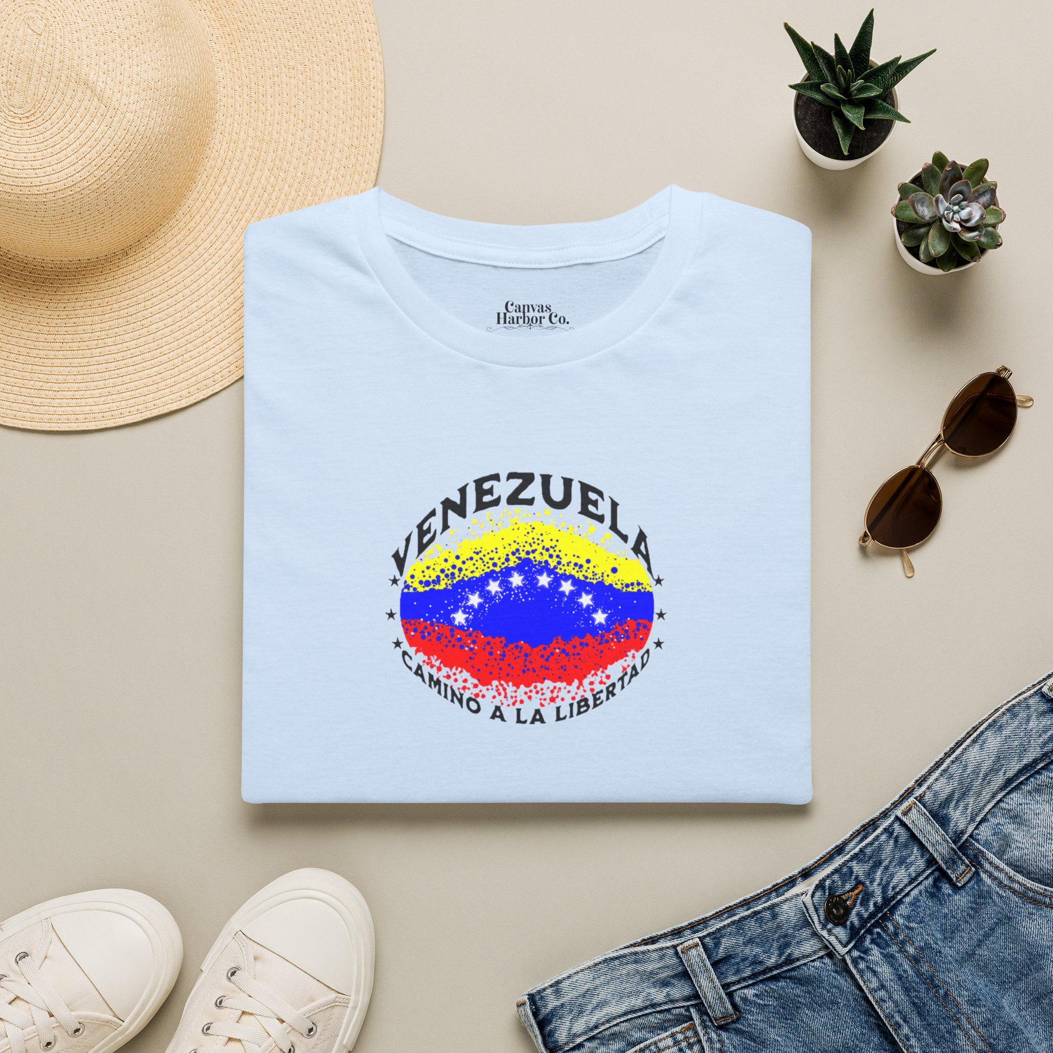 Franela azul claro, doblada en una mesa, con una bandera venezolana de forma elíptica salpicada de puntos (como una explosión), con el nombre "VENEZUELA” arriba, el texto “CAMINO A LA LIBERTAD" abajo y 3 estrellas en cada lado.