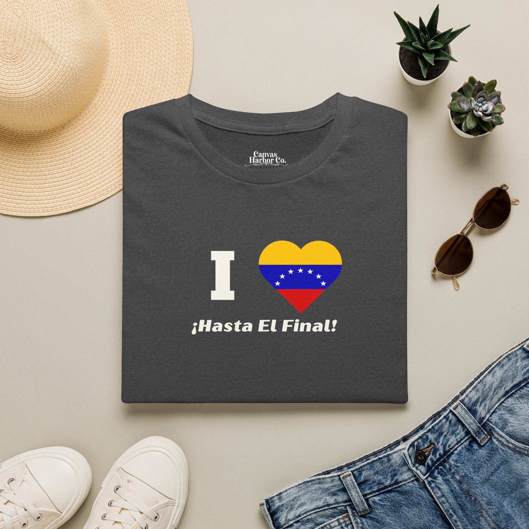 A dark gray folded T-shirt lies on a table, featuring the 7-star Venezuelan flag in the shape of a heart, the word "I" on the left, and the text "¡HASTA EL FINAL!" below.