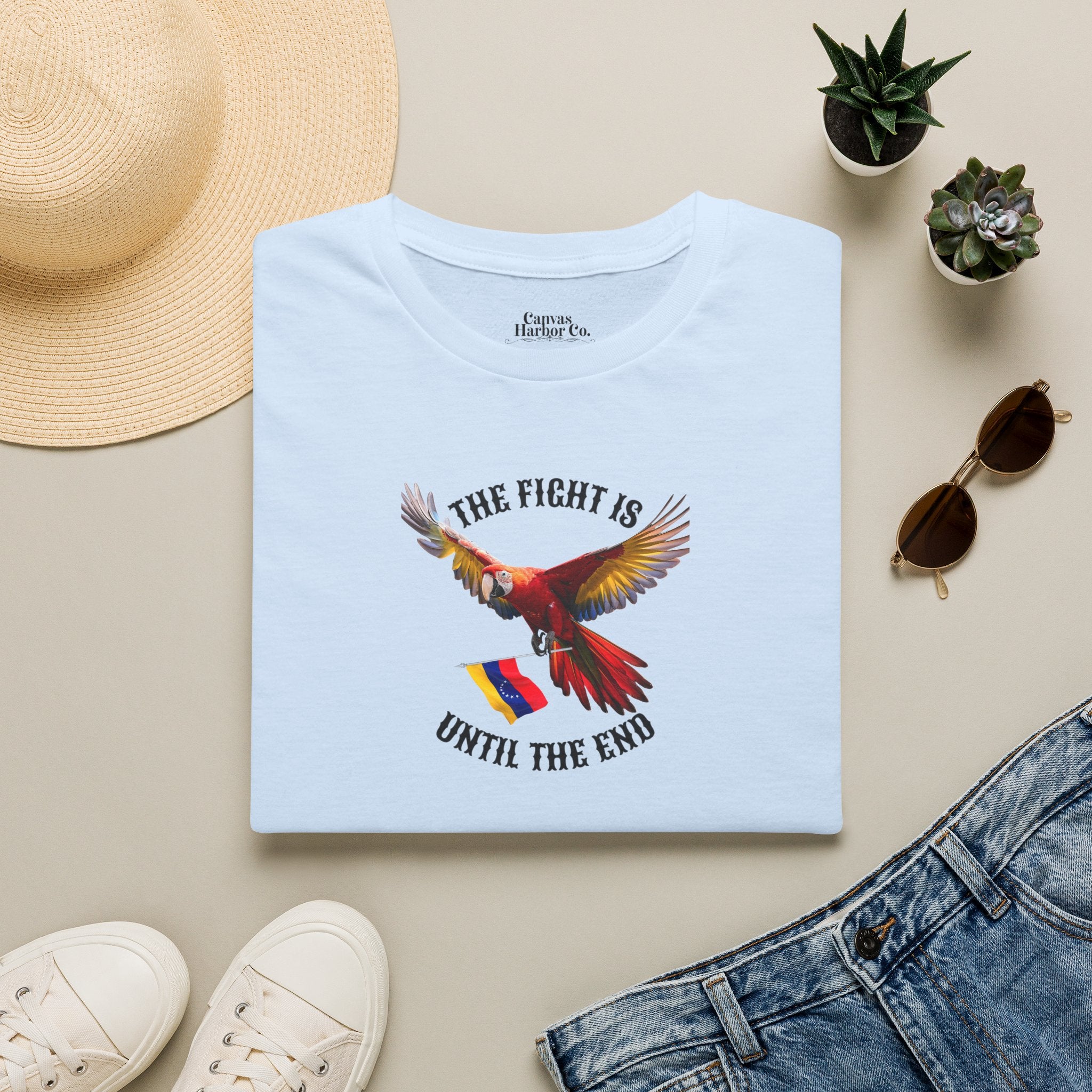 A light blue folded T-shirt lies on a table, featuring an image of a macaw and the Venezuelan flag. The text "THE FIGHT IS UNTIL THE END" surrounds the bird.