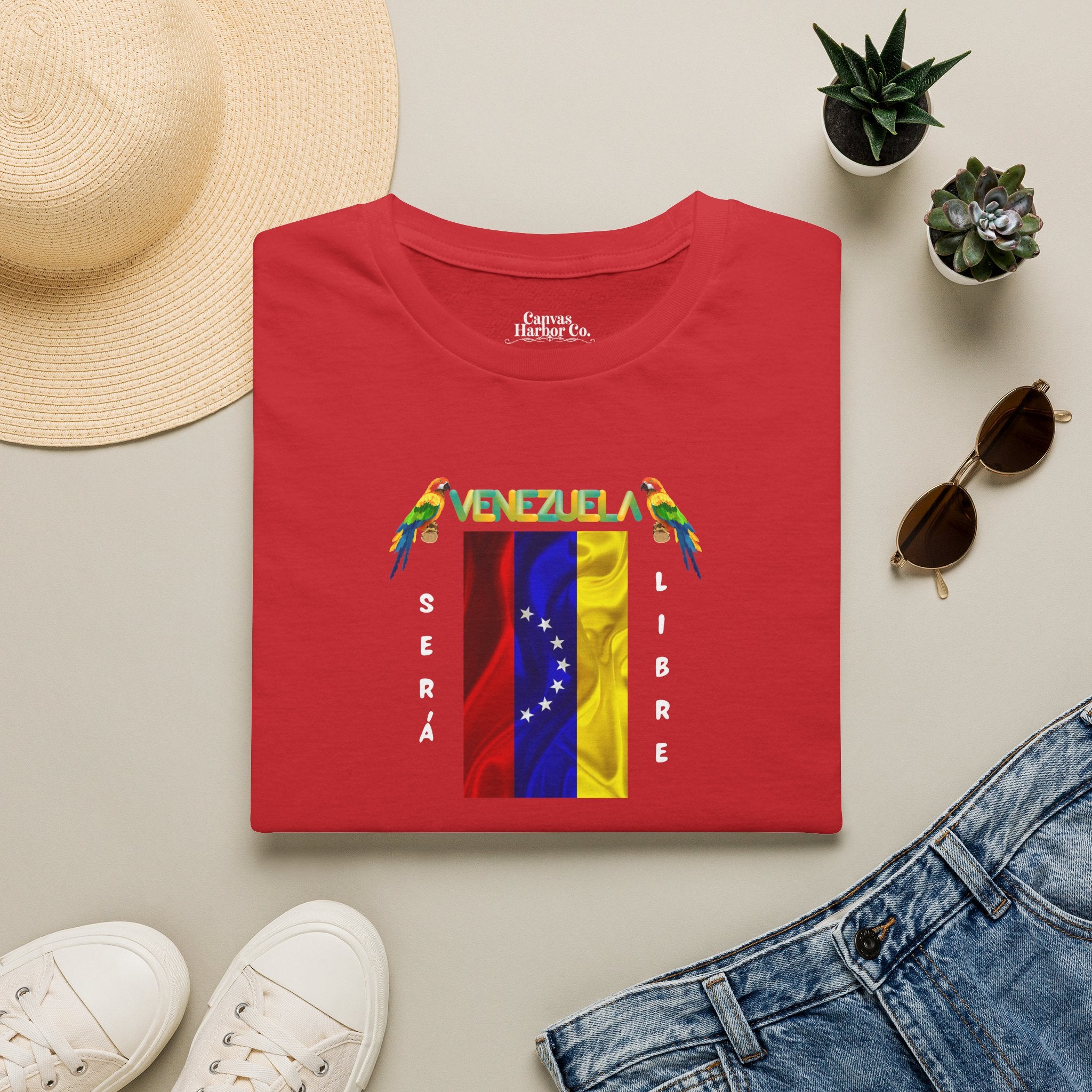Franela roja doblada sobre una mesa, con la bandera venezolana de 7 estrellas, el nombre VENEZUELA arriba y una guacamaya a cada lado. Las palabras SERÁ y LIBRE se ubican verticalmente junto a la bandera.