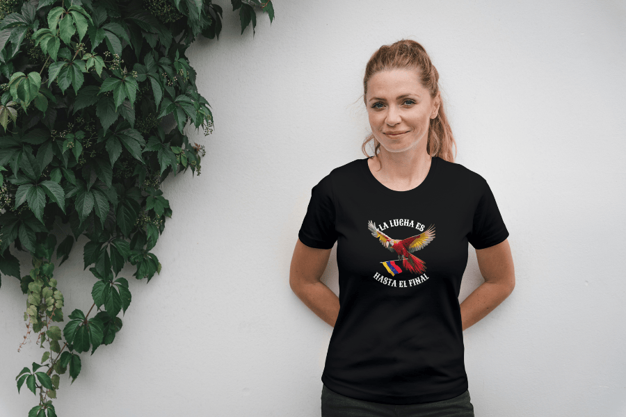 A smiling blonde woman, with both hands behind her back, wears a black unisex t-shirt featuring a colorful macaw flying while holding the Venezuelan flag. The text "La Lucha Es" is at the top, and "Hasta El Final" is below the macaw.