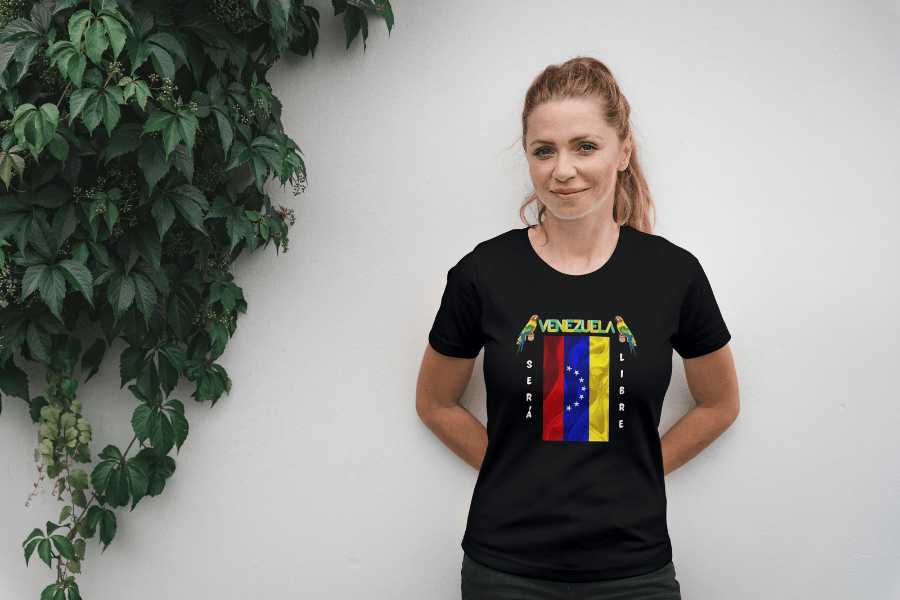 A woman wears a black unisex t-shirt featuring a Venezuelan flag, the name "Venezuela" at the top, and a colorful macaw on either side. The text "Será Libre" is vertically positioned beside the flag. She smiles, both hands behind her back.
