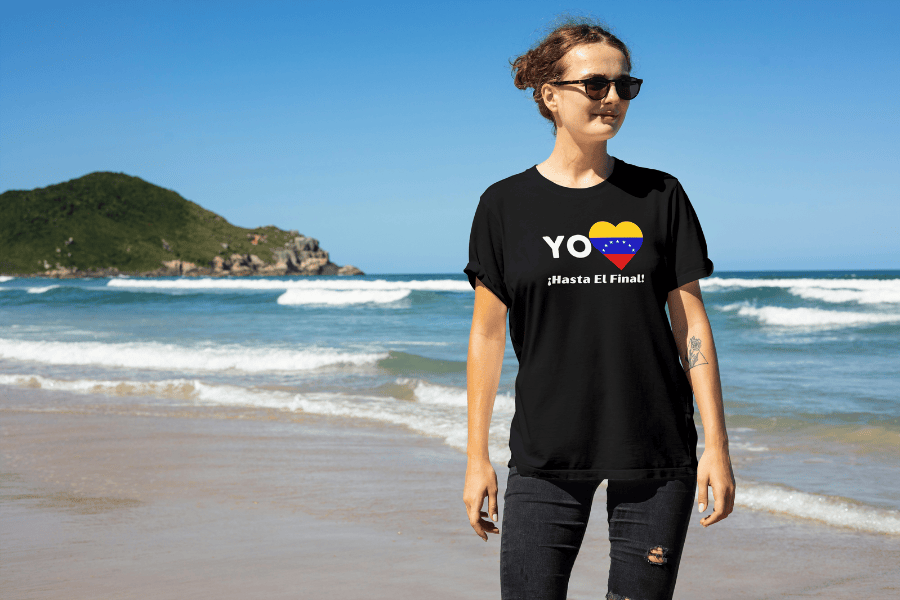 A young smiling woman wears a black unisex t-shirt featuring a heart-shaped Venezuelan flag, the word "Yo" on the left, and the text "¡Hasta El Final!" reading below. It's a sunny day; she's wearing glasses, and a beach is behind her.