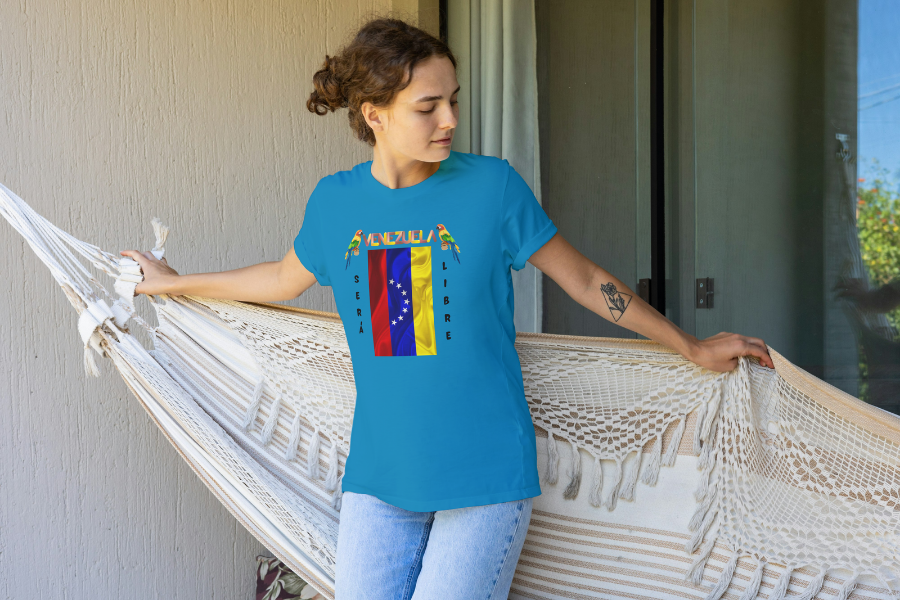 A young and beautiful woman wears a blue unisex t-shirt featuring a Venezuelan flag, the name "Venezuela" at the top, and a colorful macaw on either side. The text "Será Libre" is vertically positioned beside the flag. She's sitting on a hammock and looks down to her left.