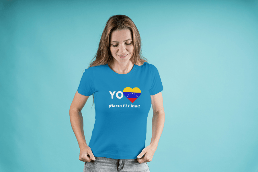 A smiling blonde woman wears a blue unisex t-shirt featuring a heart-shaped Venezuelan flag, the word "Yo" on the left, and the text “¡Hasta El Final!” below. She's looking down.