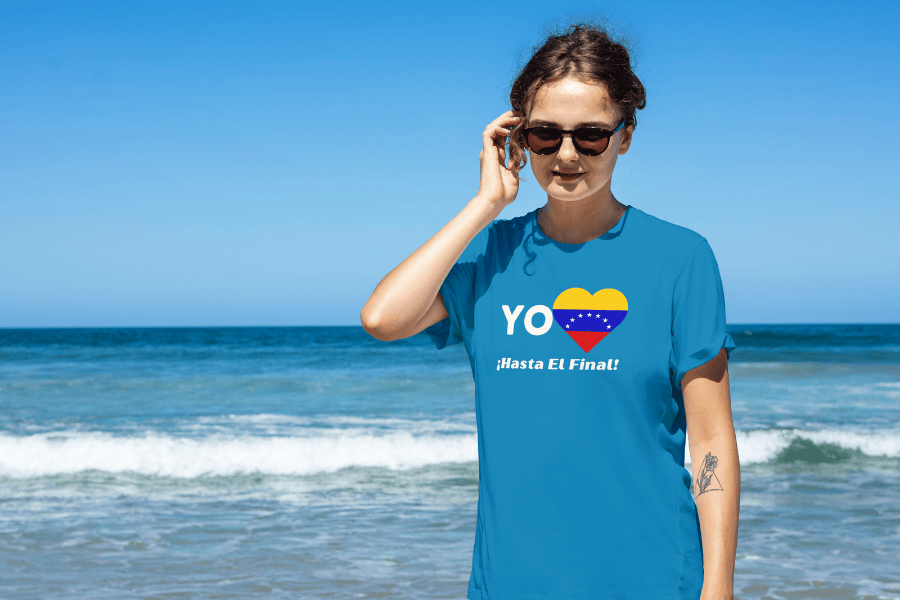 A beautiful woman wears a blue unisex t-shirt featuring a heart-shaped Venezuelan flag, the word "Yo" on the left, and the text “¡Hasta El Final!” below. It’s a sunny day, she's walking and wearing sunglasses, and a beach is behind her.