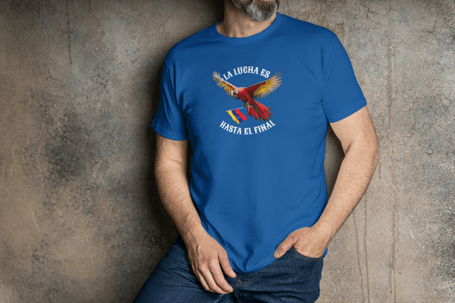 A man wears a blue unisex t-shirt featuring a colorful macaw flying while holding the Venezuelan flag. The text "La Lucha Es" is at the top, and "Hasta El Final" is below the macaw. He stands with his left hand in his pocket.