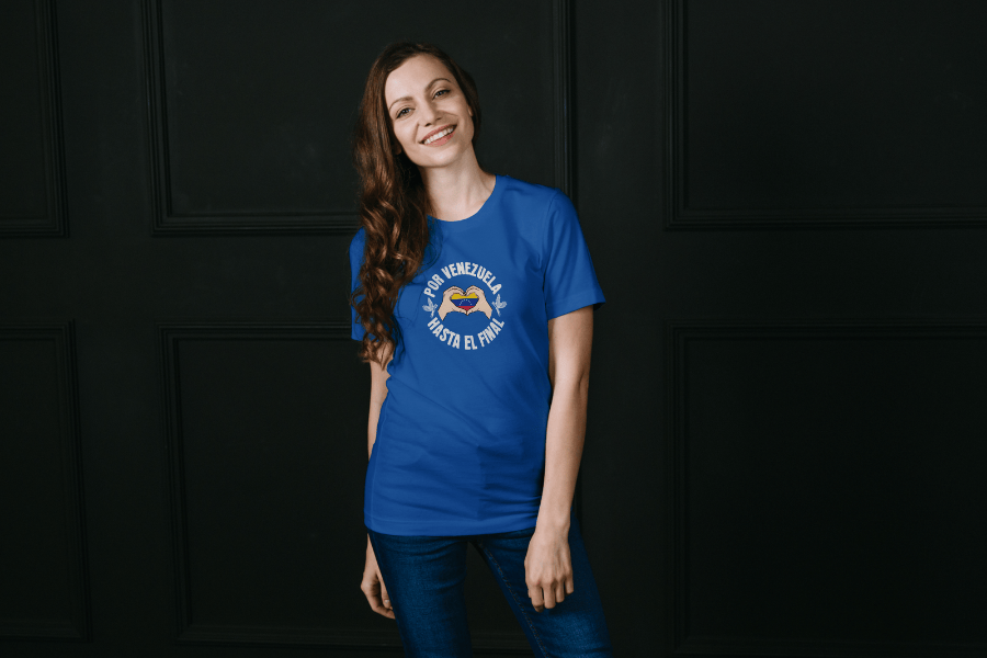 A smiling woman is wearing a blue unisex t-shirt featuring a Venezuelan flag inside heart-shaped hands, the text "Por Venezuela Hasta El Final", and a pair of macaws on either side.