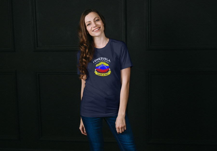A beautiful woman wears a blue unisex t-shirt featuring a colorful, elliptically shaped Venezuelan flag dotted (like an explosion), with the text "Venezuela Camino a La Libertad" at the top and "Hasta El Final" on a yellow ribbon below the flag. She’s smiling, facing forward, and a dark wall is behind her.