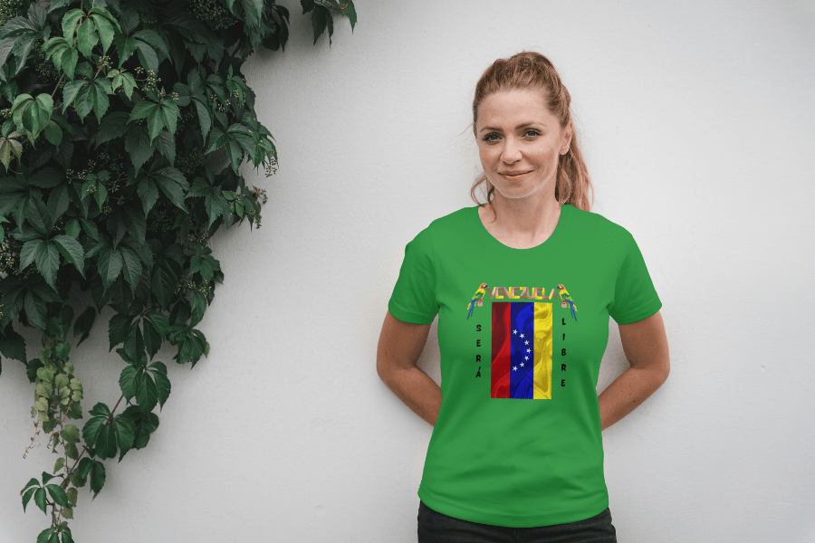A woman wears a green unisex t-shirt featuring a Venezuelan flag, the name "Venezuela" at the top, and a colorful macaw on either side. The text "Será Libre" is vertically positioned beside the flag. She smiles, both hands behind her back.