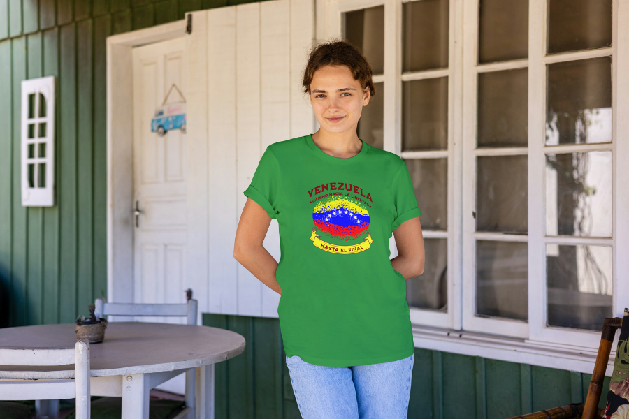 A young woman wears a green unisex t-shirt featuring a colorful, elliptically shaped Venezuelan flag dotted (like an explosion), with the text "Venezuela Camino Hacia La Libertad" at the top and "Hasta El Final" on a yellow ribbon below the flag. She's smiling, and a window is behind her.
