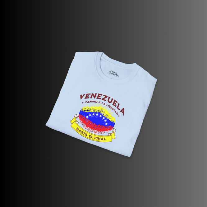 A folded light-blue unisex t-shirt featuring a colorful, elliptically shaped Venezuelan flag dotted (like an explosion), with the text "Venezuela Camino a La Libertad" at the top and "Hasta El Final" on a yellow ribbon below the flag.