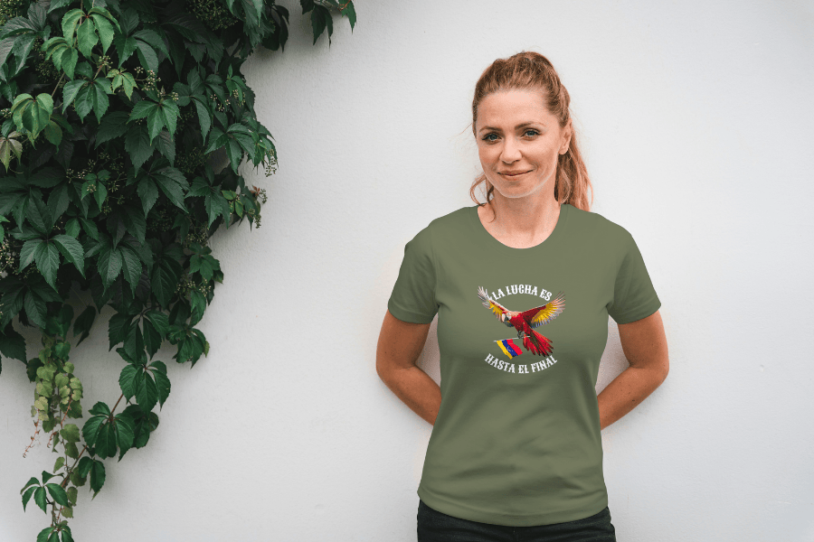 A smiling blonde woman, with both hands behind her back, wears a military green unisex t-shirt featuring a colorful macaw flying while holding the Venezuelan flag. The text "La Lucha Es" is at the top, and "Hasta El Final" is below the macaw.