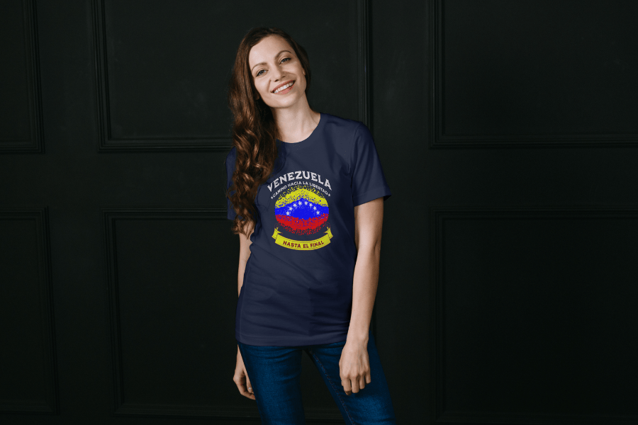 A smiling beautiful woman wears a navy unisex t-shirt featuring a colorful, elliptically shaped Venezuelan flag dotted (like an explosion), with the text "Venezuela Camino Hacia La Libertad" at the top and "Hasta El Final" on a yellow ribbon below the flag.