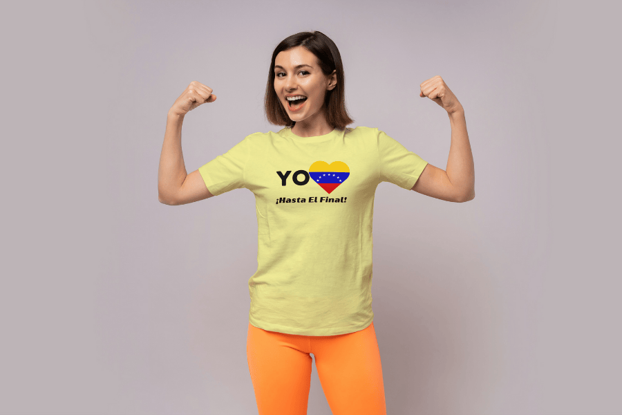 A woman is wearing a pale-yellow unisex t-shirt featuring a heart-shaped Venezuelan flag, the word "Yo" on the left, and the text "¡Hasta El Final!" reading below. She is flexing her arms and smiling.