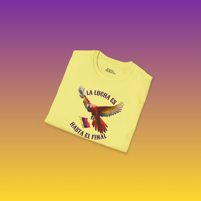 A folded pale-yellow unisex t-shirt featuring a colorful macaw flying while holding the Venezuelan flag. The text "La Lucha Es" is at the top, and "Hasta El Final" is below the macaw.
