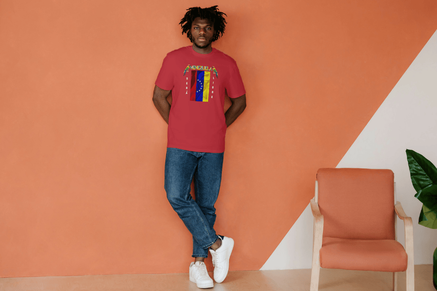 An Afro-American man wears a red unisex t-shirt featuring a Venezuelan flag, the name "Venezuela" at the top, and a colorful macaw on either side. The text "Será Libre" is vertically positioned beside the flag. He's facing forward, both hands are behind his back.
