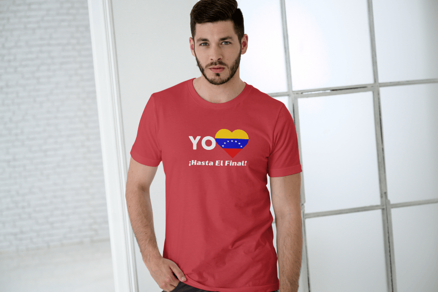 A young, bearded man is wearing a red unisex t-shirt featuring a heart-shaped Venezuelan flag, the word "Yo" on the left, and the text "¡Hasta El Final!" below. He is in a room, and a window is behind him.