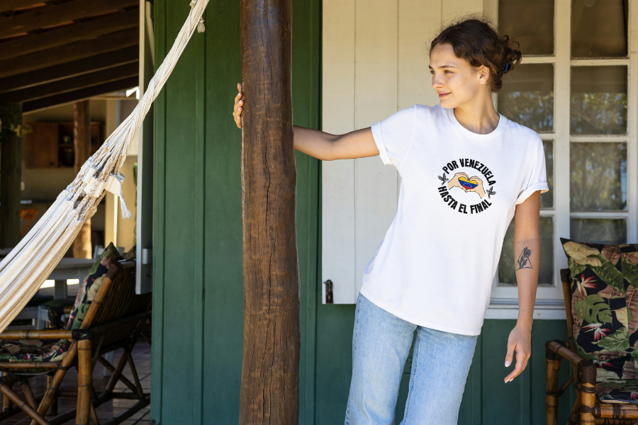A smiling woman is wearing a white unisex t-shirt featuring a Venezuelan flag inside heart-shaped hands, the text "Por Venezuela Hasta El Final", and a pair of macaws on either side. She's standing, looking to the right.