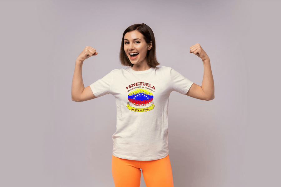 A woman wears a white unisex t-shirt featuring a colorful, elliptically shaped Venezuelan flag dotted (like an explosion), with the text "Venezuela Camino Hacia La Libertad" at the top and "Hasta El Final" on a yellow ribbon below the flag. She's flexing her arms and smiling.