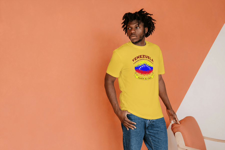 An Afro-American man is wearing a yellow unisex t-shirt featuring a colorful, elliptically Venezuelan flag dotted (like an explosion), with the text "Venezuela Camino Hacia La Libertad" at the top and "Hasta El Final" on a yellow ribbon below the flag.