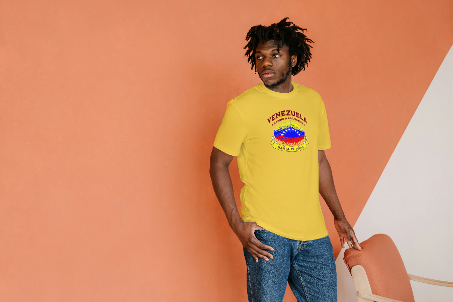 An Afro-American man is wearing a yellow unisex t-shirt featuring a colorful, elliptically shaped Venezuelan flag dotted (like an explosion), with the text "Venezuela Camino a La Libertad" at the top and "Hasta El Final" on a yellow ribbon below the flag. He’s standing, looking right.