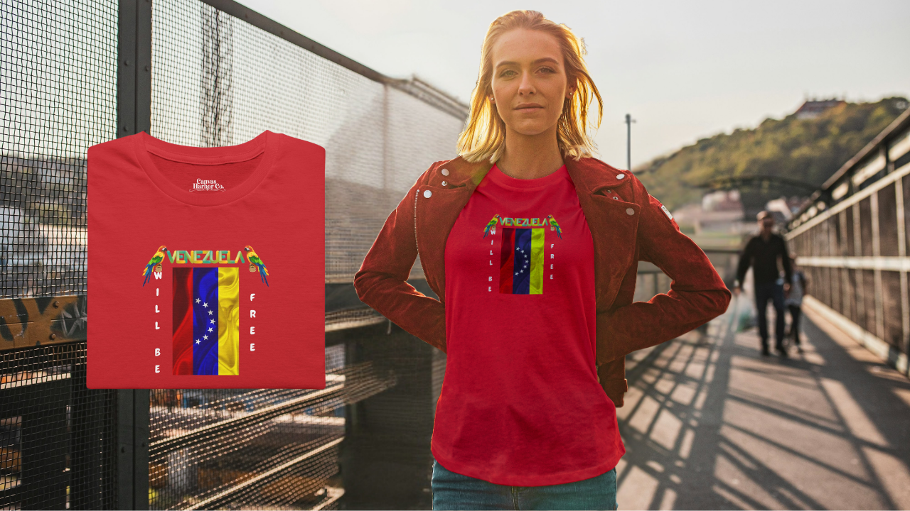 Red folded T-shirt featuring the 7-star Venezuelan flag, the name VENEZUELA at the top, and one macaw on each side. The words WILL BE and FREE are all placed vertically next to the flag. 
On the right, a young blonde woman is wearing the T-shirt. She’s standing on a bridge. 