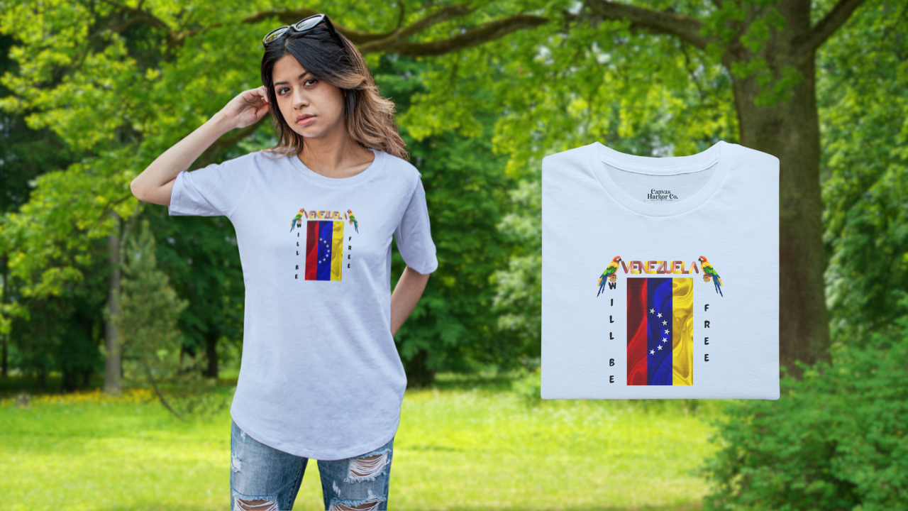 Light blue folded T-shirt featuring the 7-star Venezuelan flag, the name VENEZUELA at the top, and one macaw on each side. The words WILL BE and FREE are all placed vertically next to the flag. 
On the left, a young woman wears the T-shirt. She’s standing, facing forward, with a forest behind her.