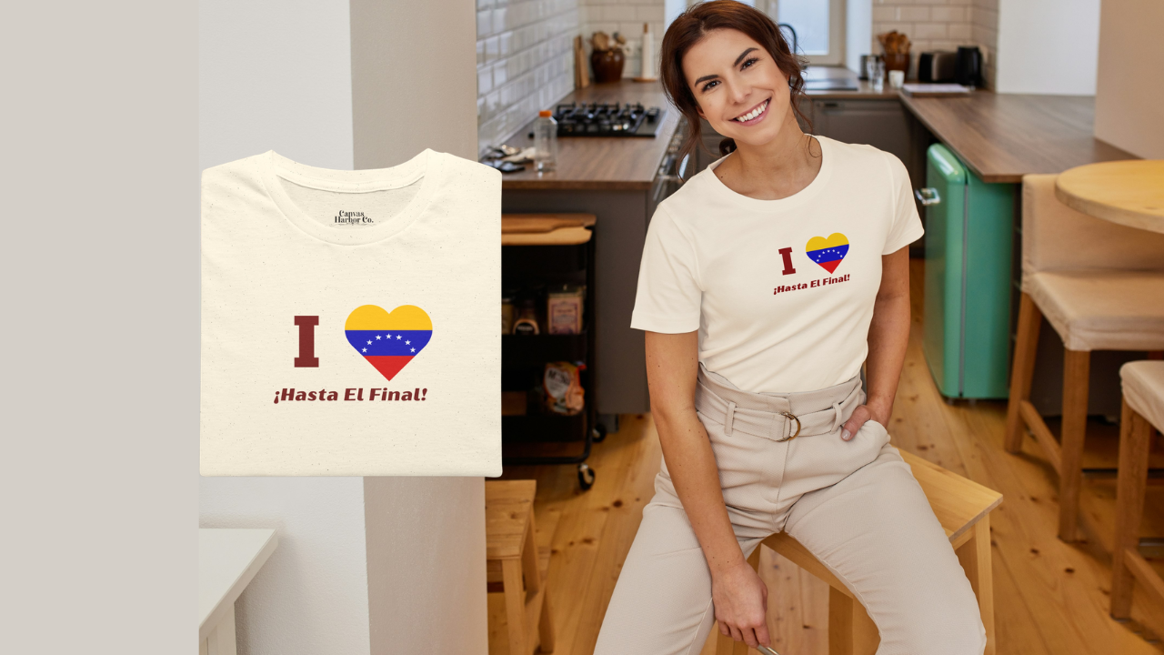 Pale orange folded T-shirt featuring the 7-star Venezuelan flag in the shape of a heart, the word "I" on the left, and the text "¡HASTA EL FINAL!" below.
On the right, a smiling woman wears the T-shirt. She’s in a kitchen, sitting on a small wooden bench.