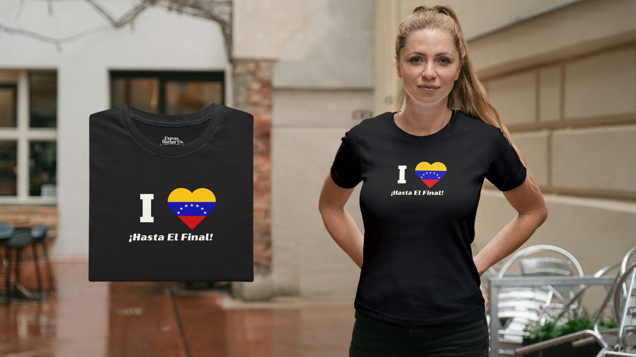 Black folded T-shirt featuring the 7-star Venezuelan flag in the shape of a heart, the word "I" on the left, and the text "¡HASTA EL FINAL!" below.
On the right, a blonde woman wears the T-shirt. She’s standing, facing forward.