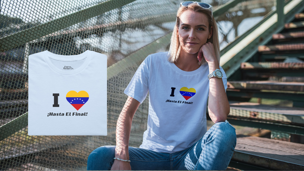 Light blue folded T-shirt featuring the 7-star Venezuelan flag in the shape of a heart, the word "I" on the left, and the text "¡HASTA EL FINAL!" below.
On the right, a blonde smiling woman wears the T-shirt. She’s facing forward, sitting on some stairs.