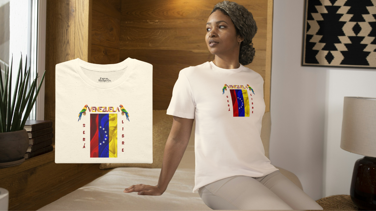 Franela doblada color crema con la bandera venezolana de 7 estrellas, el nombre VENEZUELA arriba y una guacamaya a cada lado. Las palabras SERÁ y LIBRE se ubican verticalmente junto a la bandera.
A la derecha, una mujer afroamericana viste la franela. Ella está en un cuarto, sentada sobre una cama. 