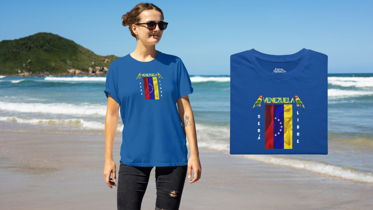 Franela doblada color azul con la bandera venezolana de 7 estrellas, el nombre VENEZUELA arriba y una guacamaya a cada lado. Las palabras SERÁ y LIBRE se ubican verticalmente junto a la bandera.
A la izquierda, hay una mujer sonriente, en una playa, vistiendo la franela. 