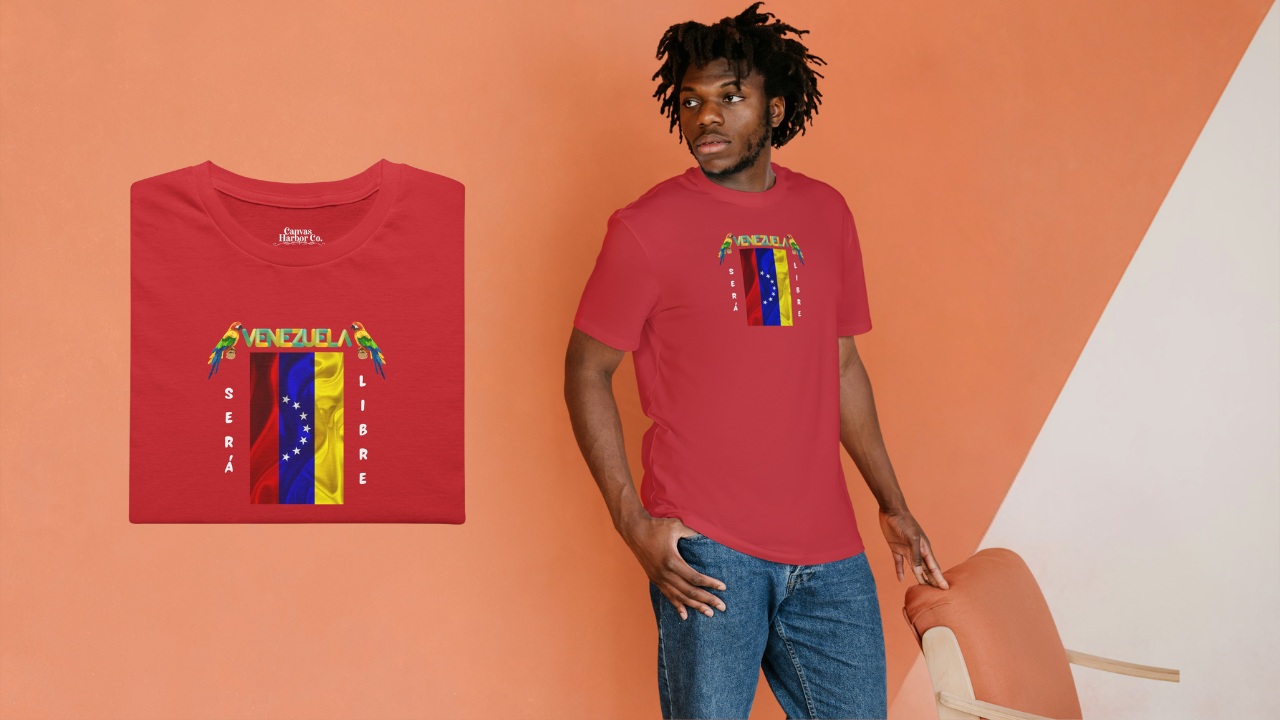 Franela doblada color rojo con la bandera venezolana de 7 estrellas, el nombre VENEZUELA arriba y una guacamaya a cada lado. Las palabras SERÁ y LIBRE se ubican verticalmente junto a la bandera.
A la derecha, hay un hombre afroamericano de pie vistiendo la franela. 