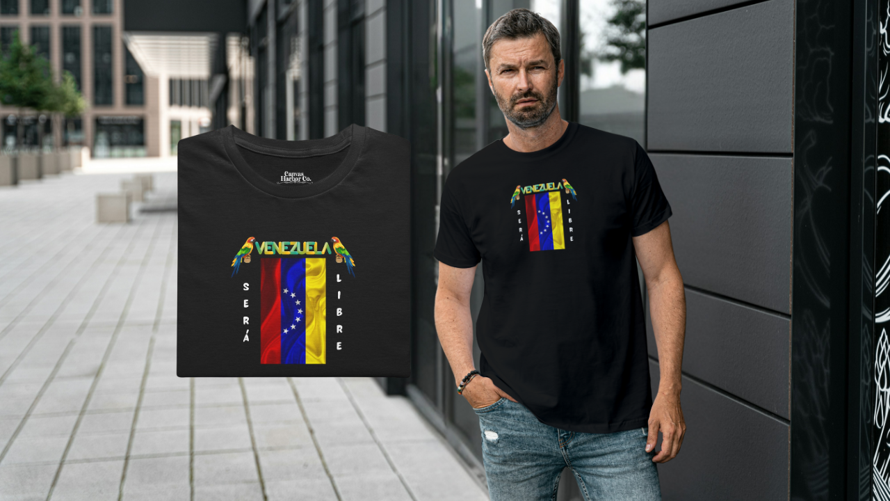 Franela doblada color negro con la bandera venezolana de 7 estrellas, el nombre VENEZUELA arriba y una guacamaya a cada lado. Las palabras SERÁ y LIBRE se ubican verticalmente junto a la bandera.
A la derecha, un hombre con barba viste la franela. Él está de pie, frente a la entrada de un edificio. 