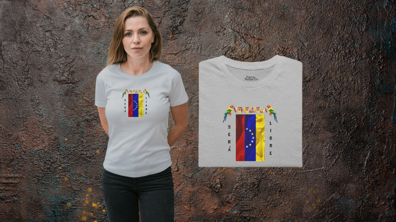 Franela doblada color gris claro con la bandera venezolana de 7 estrellas, el nombre VENEZUELA arriba y una guacamaya a cada lado. Las palabras SERÁ y LIBRE se ubican verticalmente junto a la bandera.
A la izquierda, hay una mujer joven que viste la franela. Ella está de pie, con ambas manos detrás de su espalda. 