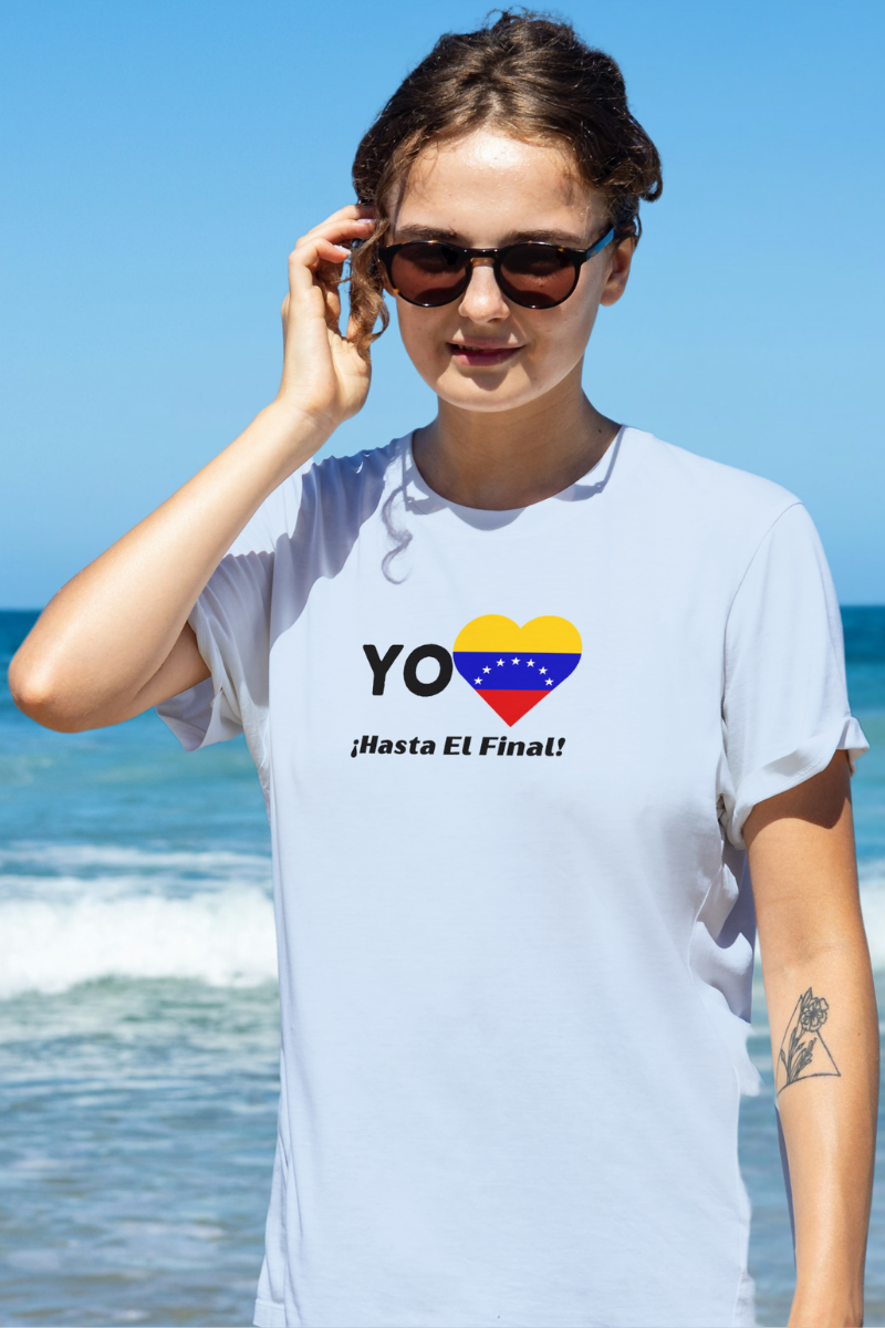 Una mujer joven viste una franela azul claro con la bandera venezolana de 7 estrellas en forma de corazón, la palabra "YO" a la izquierda y el texto "¡HASTA EL FINAL!" debajo.
Ella está en una playa, usando lentes de sol.