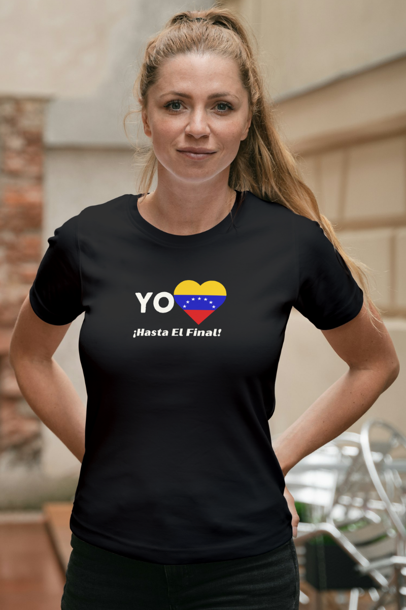 Una mujer joven y rubia viste una franela negra con la bandera venezolana de 7 estrellas en forma de corazón, la palabra "YO" a la izquierda y el texto "¡HASTA EL FINAL!" debajo.
Ella está de pie, mirando al frente.