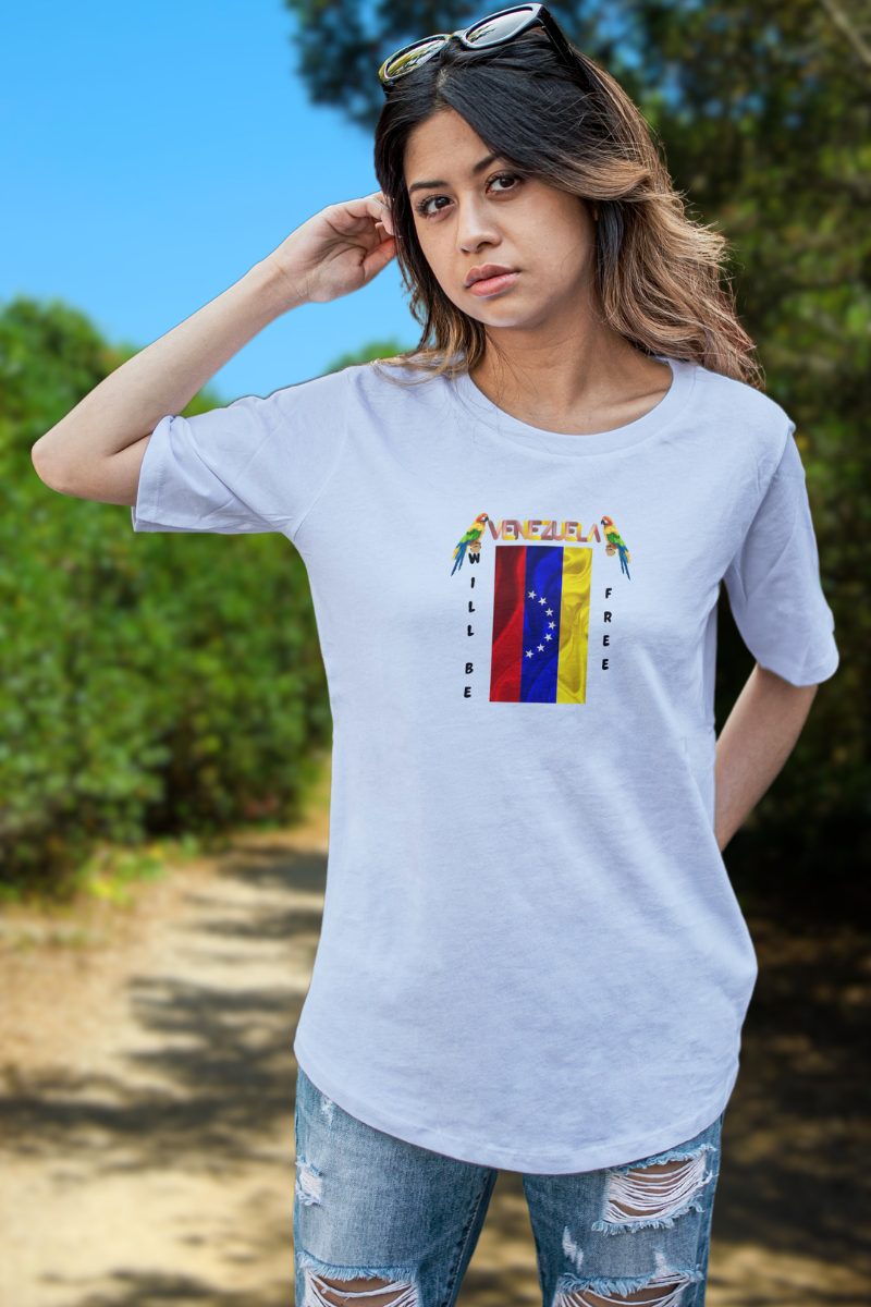 A young woman wears a light blue T-shirt featuring the 7-star Venezuelan flag, the name VENEZUELA at the top, and one macaw on each side. The words WILL BE and FREE are all placed vertically next to the flag. 
She’s standing, facing forward, with a forest behind her.
