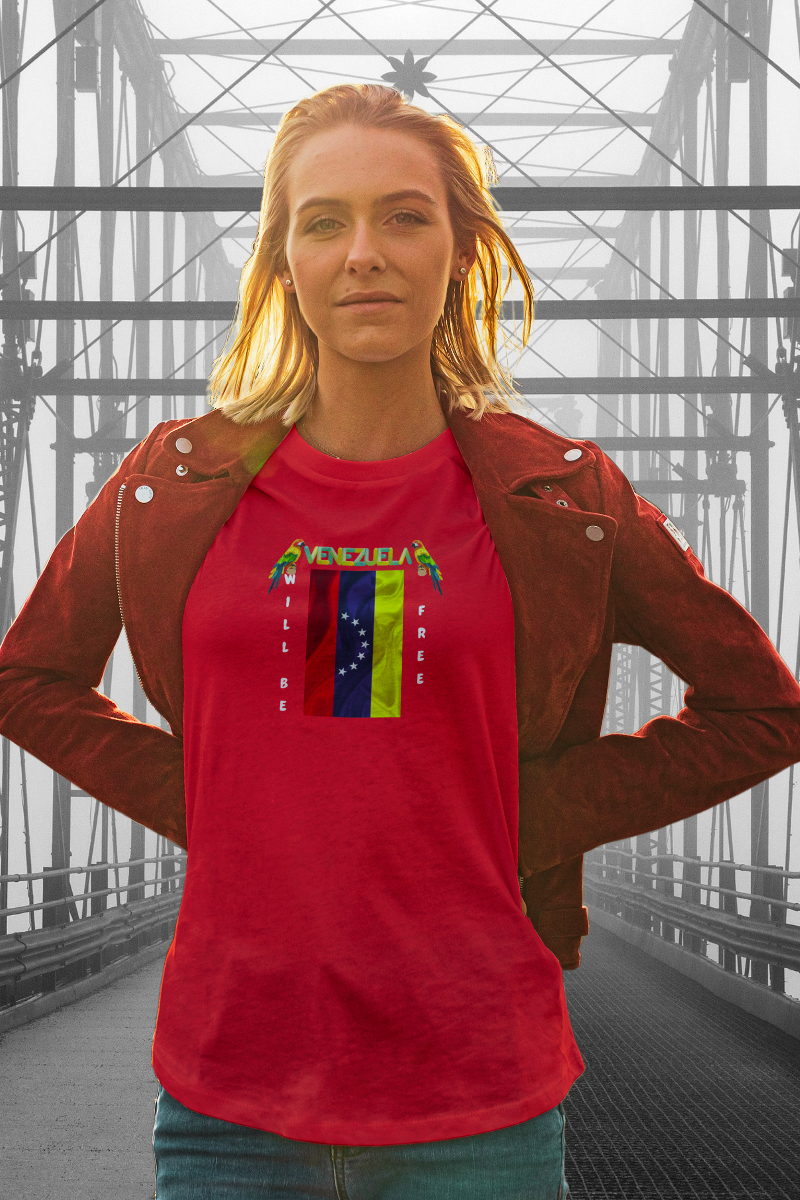 A young blonde woman is wearing a red T-shirt featuring the 7-star Venezuelan flag, the name VENEZUELA at the top, and one macaw on each side. The words WILL BE and FREE are all placed vertically next to the flag. 
She’s standing on a bridge. 