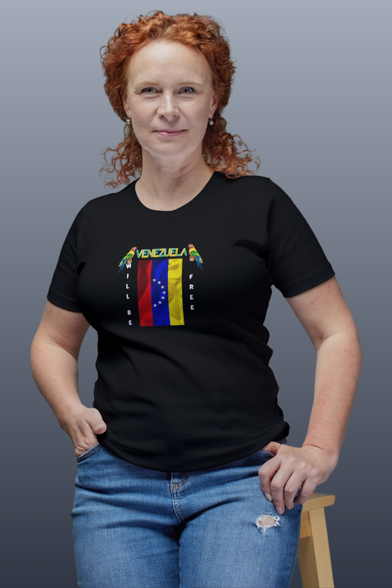 A red-haired woman is wearing a black T-shirt featuring the 7-star Venezuelan flag, the name VENEZUELA at the top, and one macaw on each side. The words WILL BE and FREE are all placed vertically next to the flag. 
She’s smiling, sitting on a small wooden bench. 